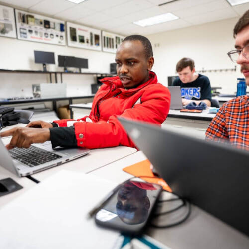 Two students in a classroom work together on laptops, with one in a red jacket pointing at the screen while the other observes, surrounded by electronic equipment and other focused classmates.